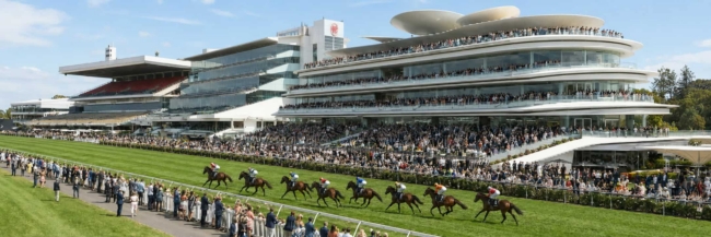 Grandstand filled with spectators at a horse race as horses run on a sunny track, with a modern multi-tiered venue in the background.