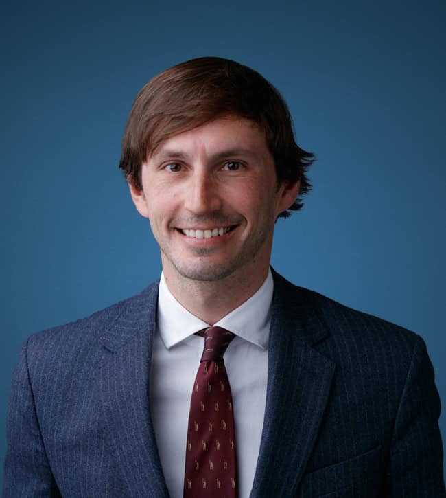 Smiling man in a navy pinstripe suit and burgundy tie against a blue backdrop, head-and-shoulders portrait.
